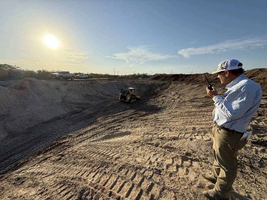 A construction worker wearing a hard hat stands in a large dirt construction site, observing a small bulldozer at work in the distance. The scene shows an active land development project with rolling hills, scattered trees, and wind turbines visible on the horizon.