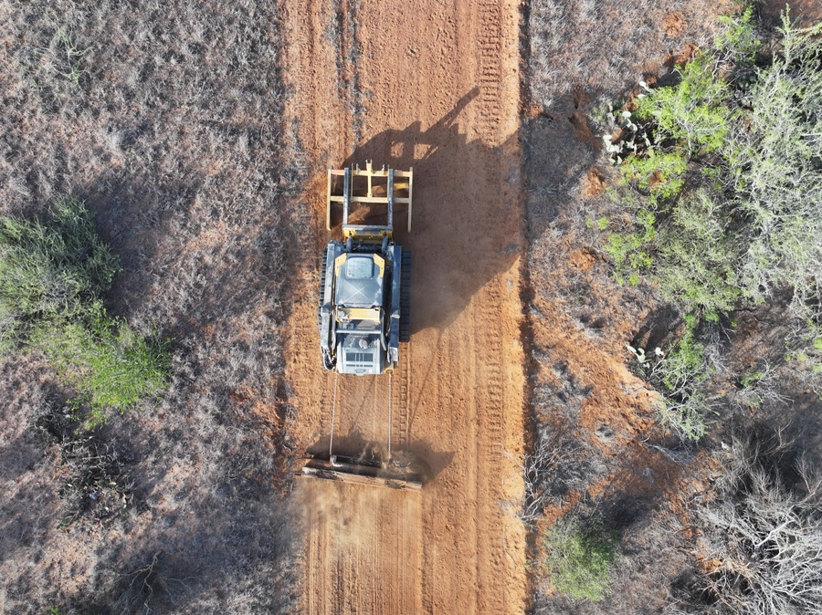 Aerial view of a large yellow construction vehicle excavating and moving dirt on a rural, unpaved road through a wooded area.