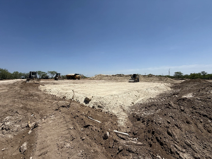 A large, open construction site with heavy equipment including bulldozers and dump trucks moving dirt and clearing the land for a new development project.