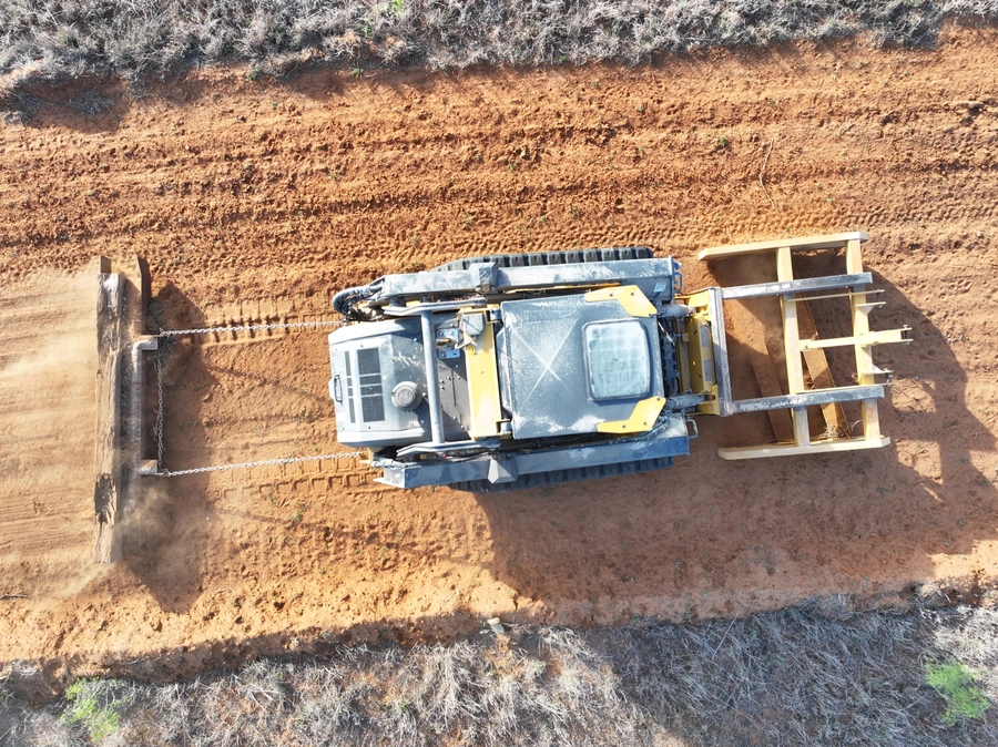 An aerial view of a construction vehicle, likely a backhoe or excavator, working on a dirt and gravel construction site surrounded by shrubs and vegetation.