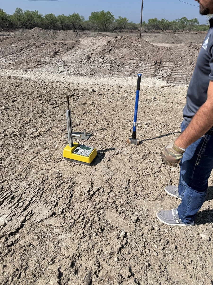 A construction site with heavy machinery, tools, and a surveyor's equipment. Dirt and debris cover the ground, indicating ongoing excavation or land preparation work.