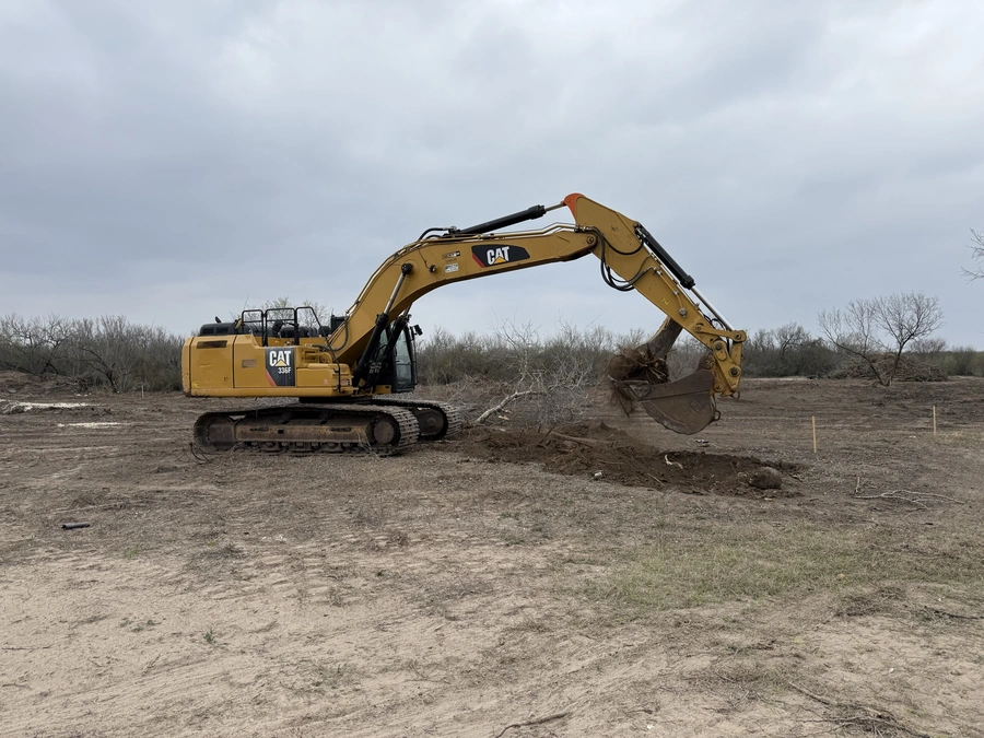 A large yellow Caterpillar excavator with a bucket attachment digging into the dirt at a construction site. Bare trees and a gray cloudy sky can be seen in the background.