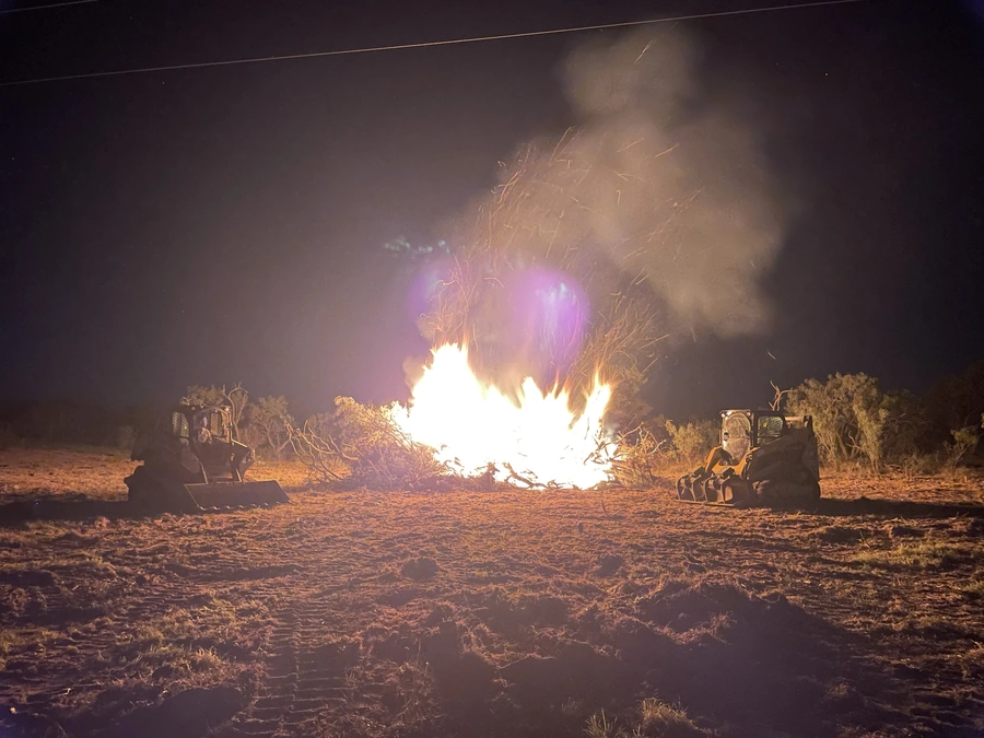A nighttime scene showing a large controlled burn on a rural landscape. A bulldozer and other heavy machinery are visible as bright flames and smoke billow into the dark sky.
