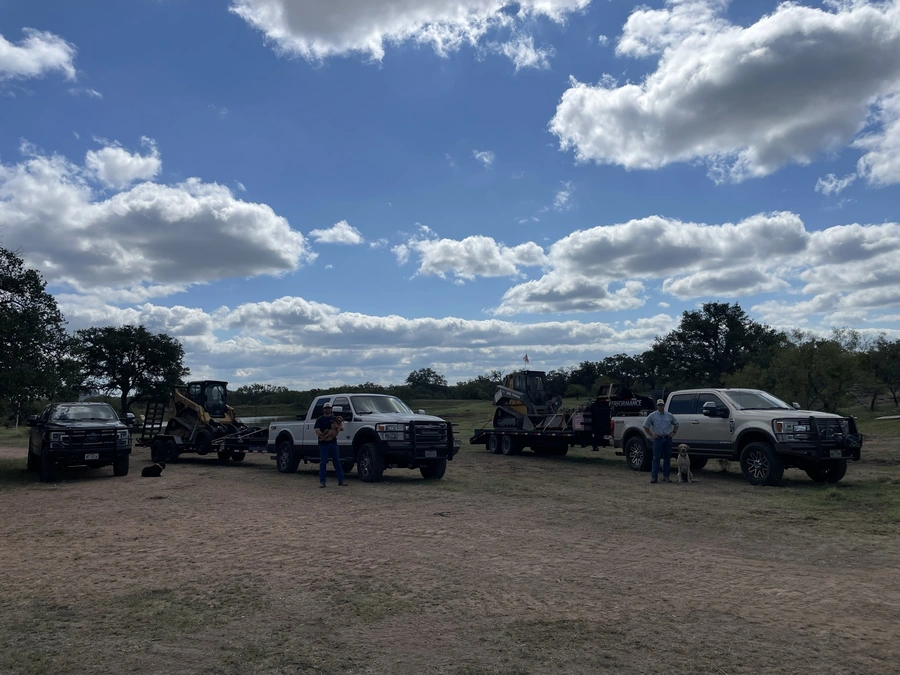 A scene showing various construction vehicles and equipment, including pickup trucks, a dump truck, and a backhoe, set against a scenic landscape with a blue sky and puffy white clouds. Several people are visible, including one standing in the foreground.