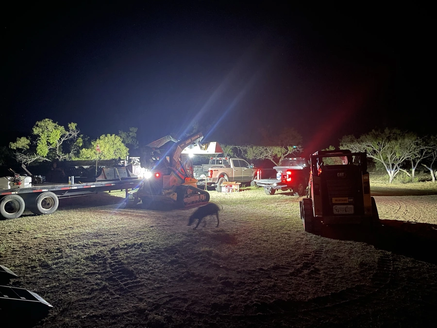 A night construction site with heavy machinery, trucks, and equipment illuminated by bright lights. The scene depicts the active work being done on a project, possibly at a ranch or rural location.