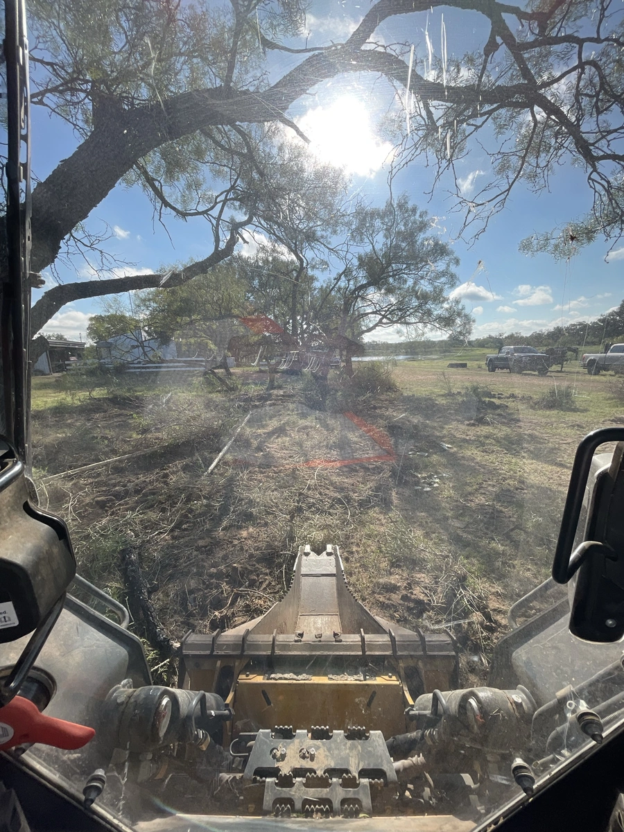 View from the cab of a bulldozer or other heavy equipment on a ranch or construction site. The image shows a cleared path through the brush and trees, with a blue sky and scattered clouds visible overhead.