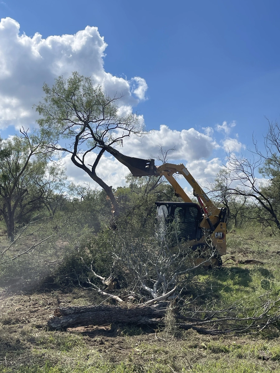 A heavy machinery excavator is removing fallen tree branches and debris from a wooded area on a ranch. The excavator's arm is extended, grasping a large tree branch that has been cut or broken off. The sky is filled with fluffy white clouds against a bright blue background.