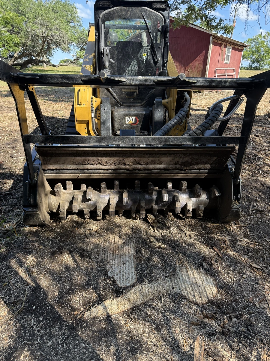 An image of a yellow Caterpillar skid steer loader with a toothed bucket attachment, operating in a rural setting with a red barn in the background.