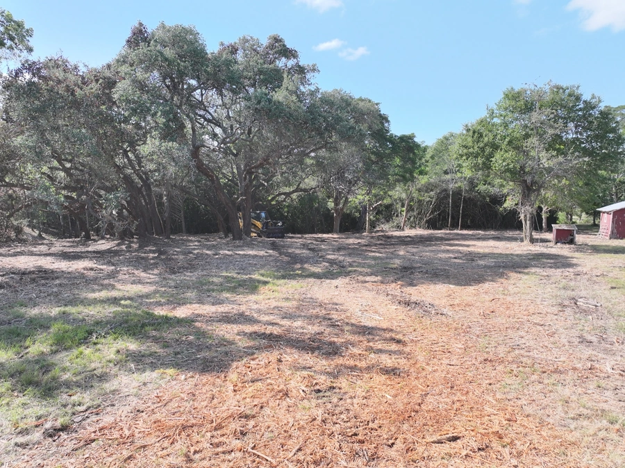 A cleared, dirt construction site surrounded by lush green trees. A backhoe vehicle is visible in the background, indicating ongoing land clearing or site preparation work.