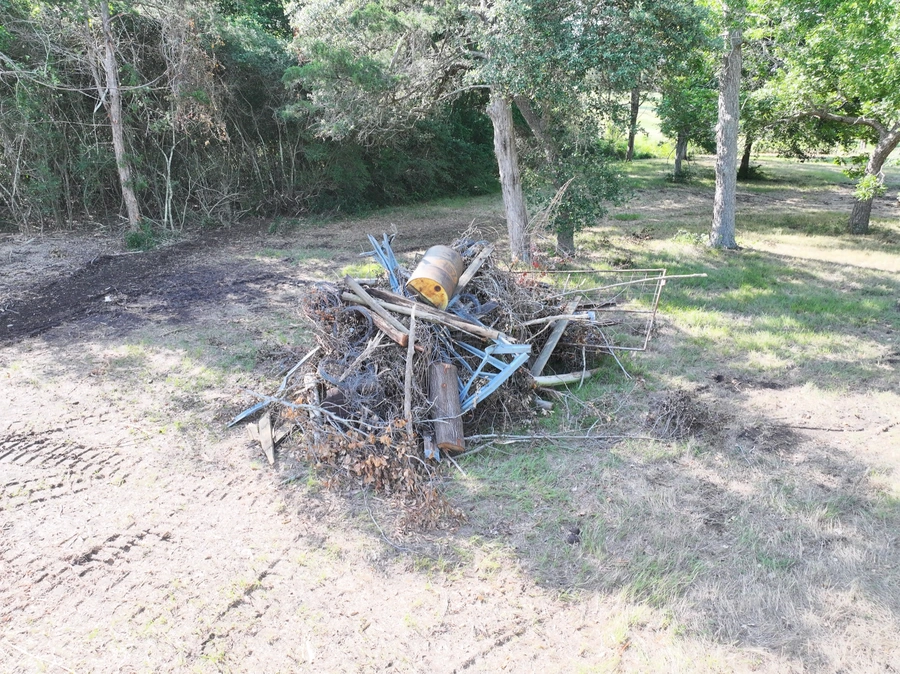 A large pile of discarded and damaged construction materials and debris, including metal pieces, broken wood, and other waste, lying in a grassy clearing surrounded by trees in a wooded area.