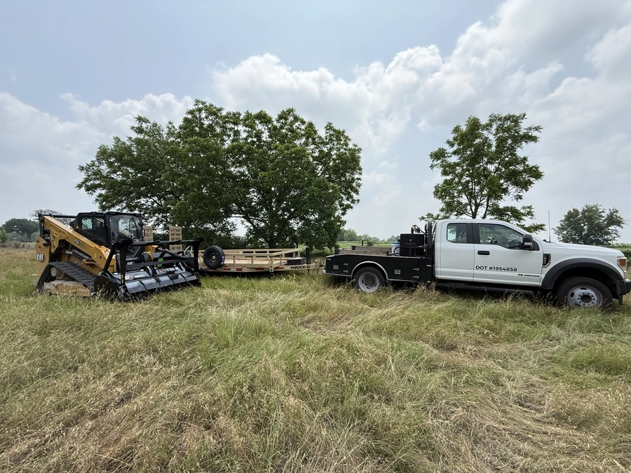 Construction equipment, including a Caterpillar skid steer loader and a truck, parked in a grassy field with trees in the background. The scene suggests land improvement or site work being carried out by a construction company.