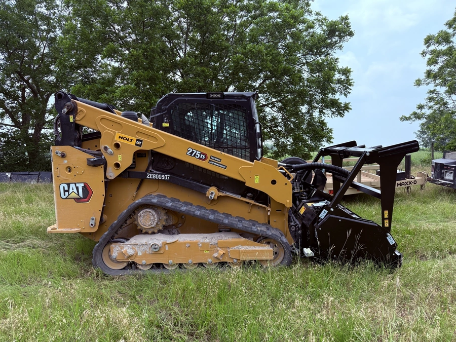 A yellow Caterpillar brand skid-steer loader with a black attachment on a grassy field surrounded by trees. The skid-steer loader appears to be used for construction or land clearing work.