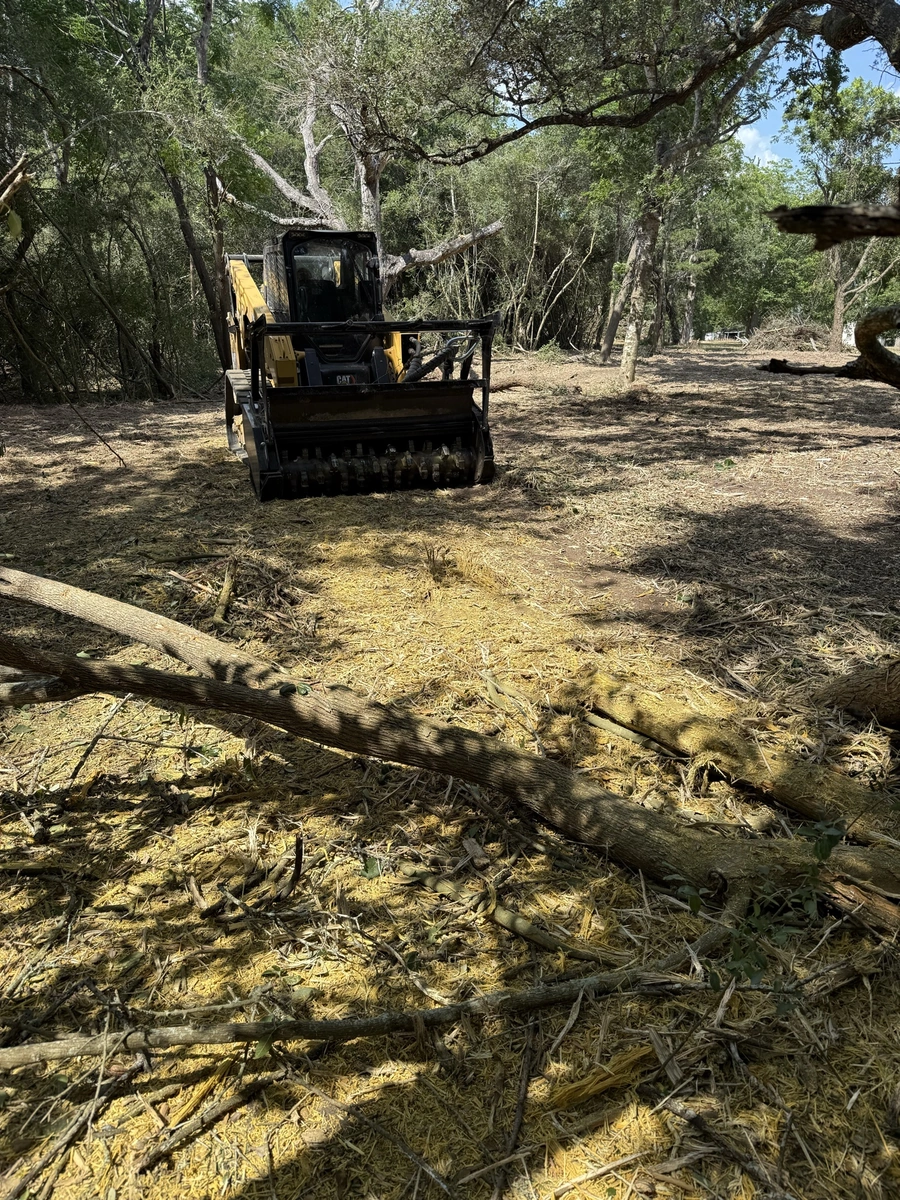 A yellow backhoe or skid steer loader clearing debris and vegetation in a wooded area, with fallen branches and trees visible on the ground.