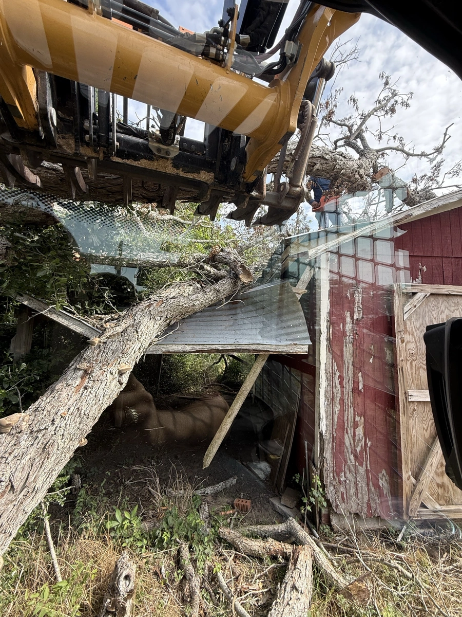 A large construction vehicle with a raised bucket or arm is shown in the foreground, surrounded by debris and a partially damaged wooden structure or shed in the background. The image depicts the aftermath of a destructive incident, likely related to land clearing, excavation, or storm damage.