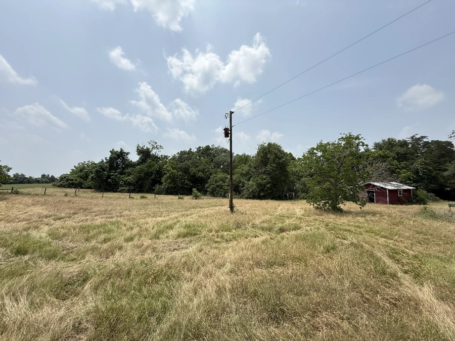 A rural field with a red barn in the distance, surrounded by lush green trees. Power lines and a utility pole can be seen in the foreground, indicating the presence of infrastructure in the area.