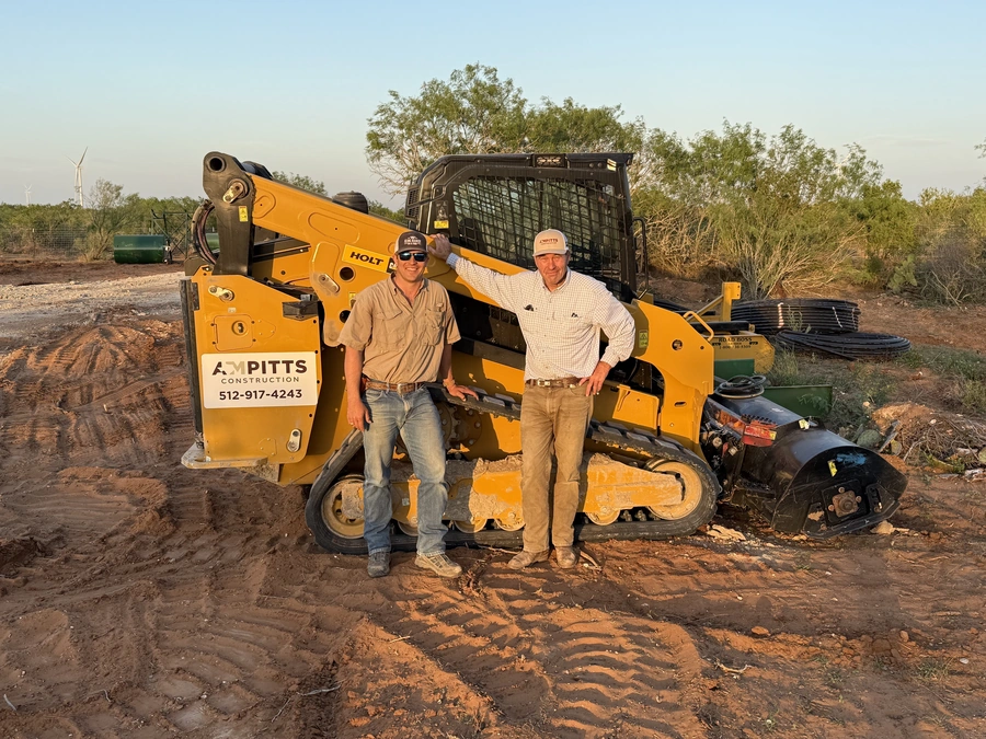 Mason and his father standing next to a yellow Holt skid-steer loader with A&M Pitts Construction branding, photographed at a construction site during golden hour with trees and wind turbines in the background