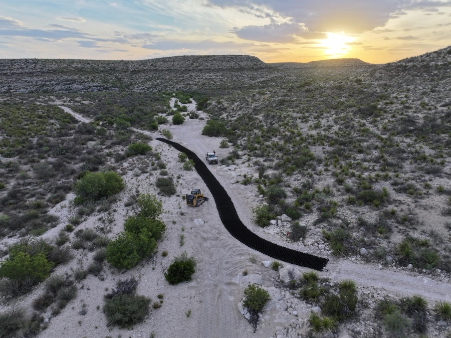 Aerial view of a dirt road winding through a rugged, arid landscape at sunset. In the foreground, construction equipment can be seen working on the road.