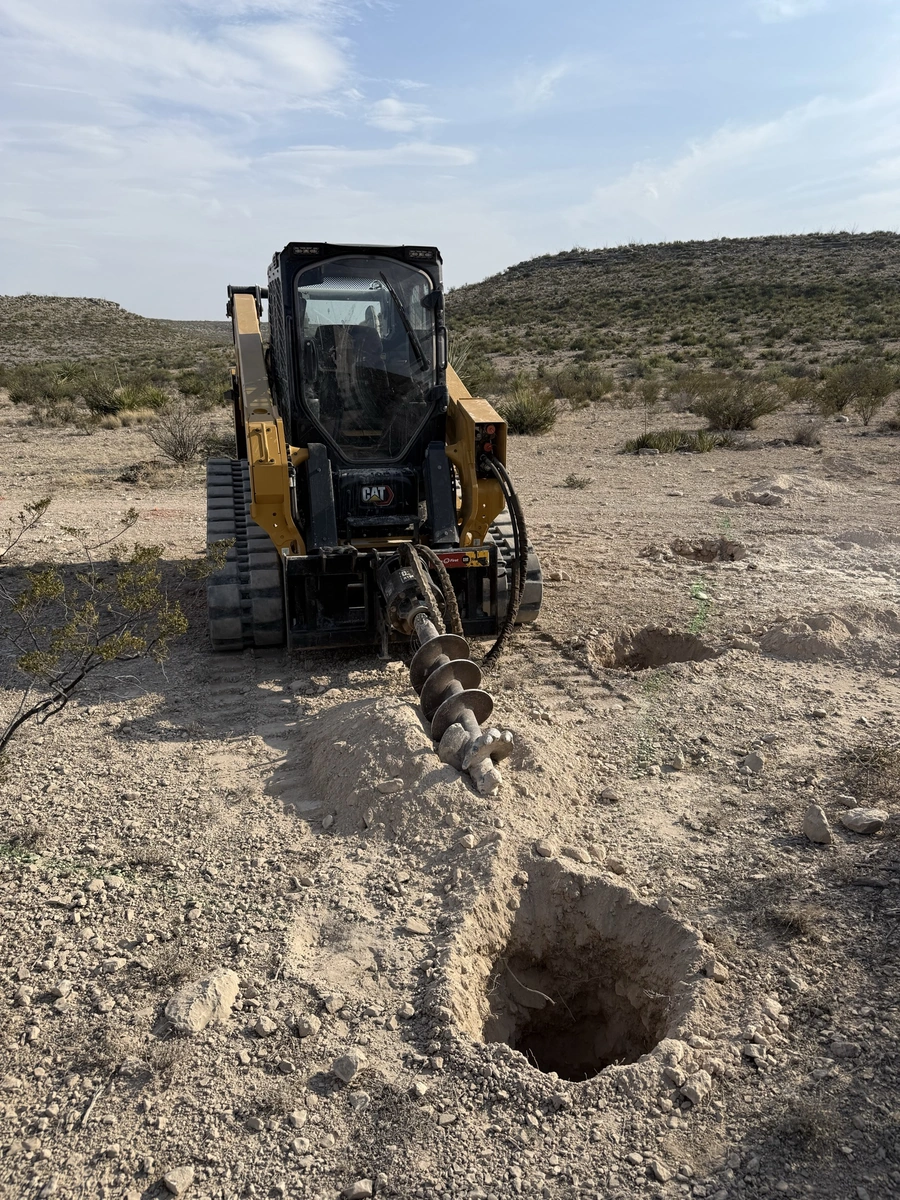 A Caterpillar skid steer loader is digging a trench in a desert landscape with mountains in the background under a blue cloudy sky.