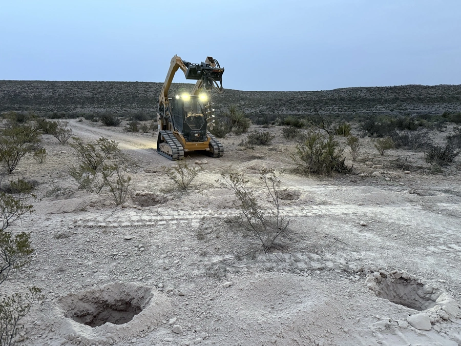 A large yellow excavator machine operating in a remote desert landscape at night, with its powerful headlights illuminating the rugged terrain.
