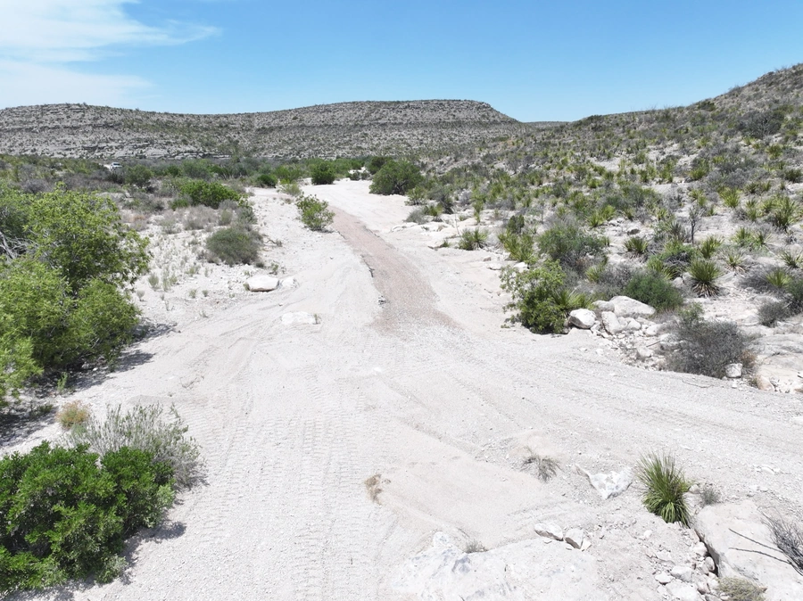 A winding unpaved road stretches through a rugged desert landscape, surrounded by hills and vegetation. The scene conveys the remote, off-the-beaten-path nature of the location.