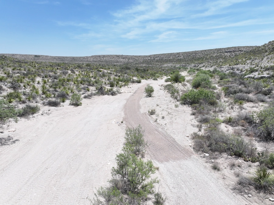 A remote, rugged desert landscape with a dirt road winding through the dry, rocky terrain and sparse vegetation. The sky is a bright, clear blue with scattered clouds.