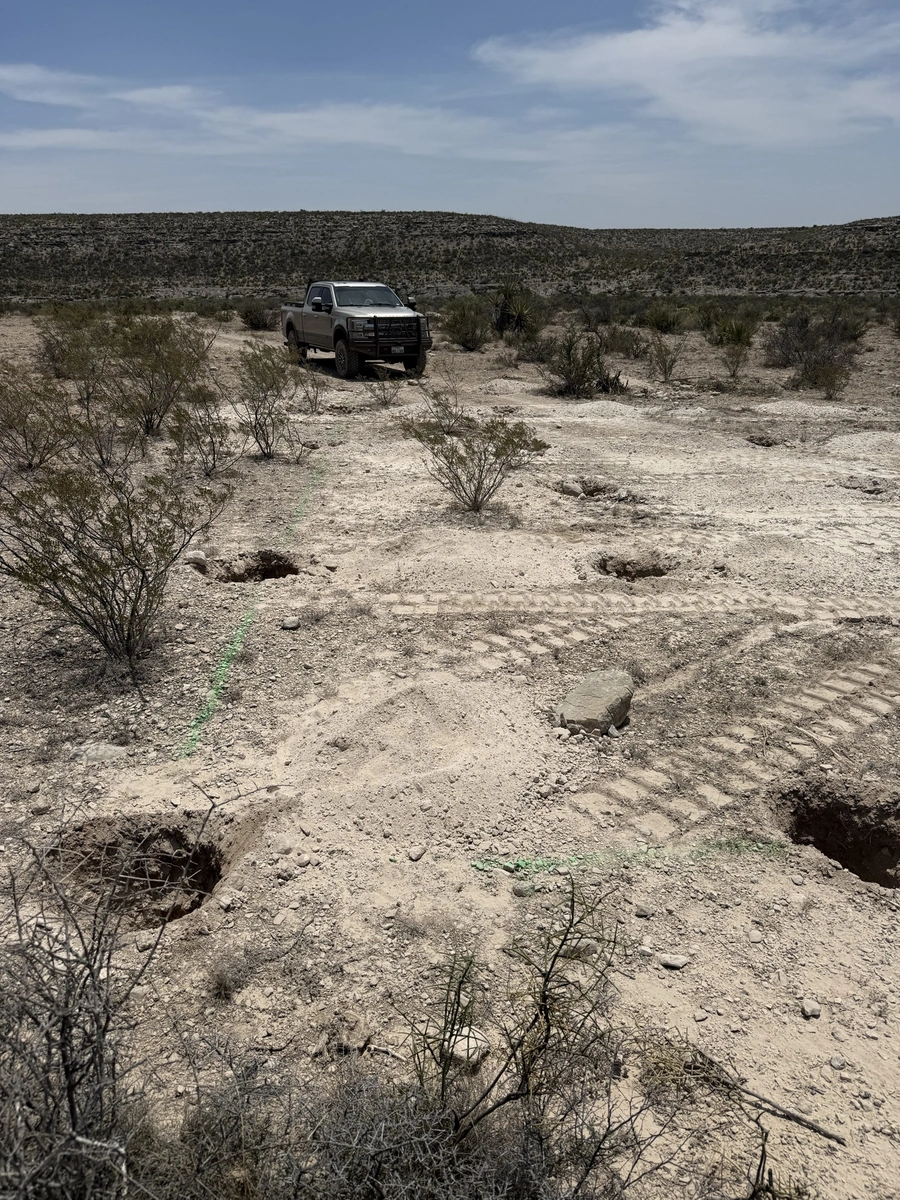 A rugged pickup truck navigates a remote, rocky desert landscape under a bright blue sky with scattered clouds. The terrain is sparse, with scattered shrubs and vegetation, suggesting a challenging off-road environment.