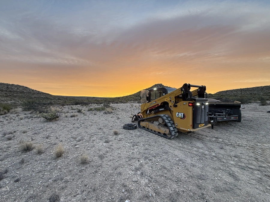 A Caterpillar excavator or skid steer loader sits in a desert landscape at sunset, with mountains in the background and a vibrant orange and purple sky.