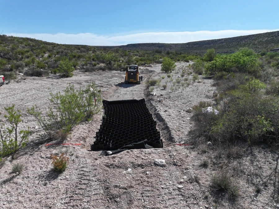A bulldozer moving along a dirt road in a rugged, mountainous desert landscape with sparse vegetation. The image suggests the type of terrain and land improvement work that A.M. Pitts Construction specializes in.