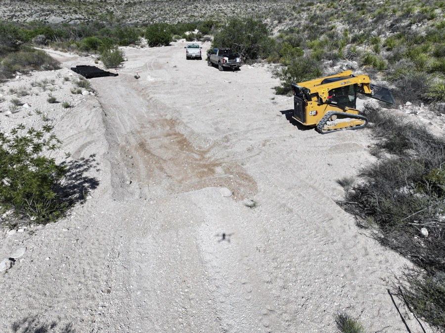A Caterpillar excavator machine working on a dirt road in a remote, arid landscape with shrubs and vegetation.