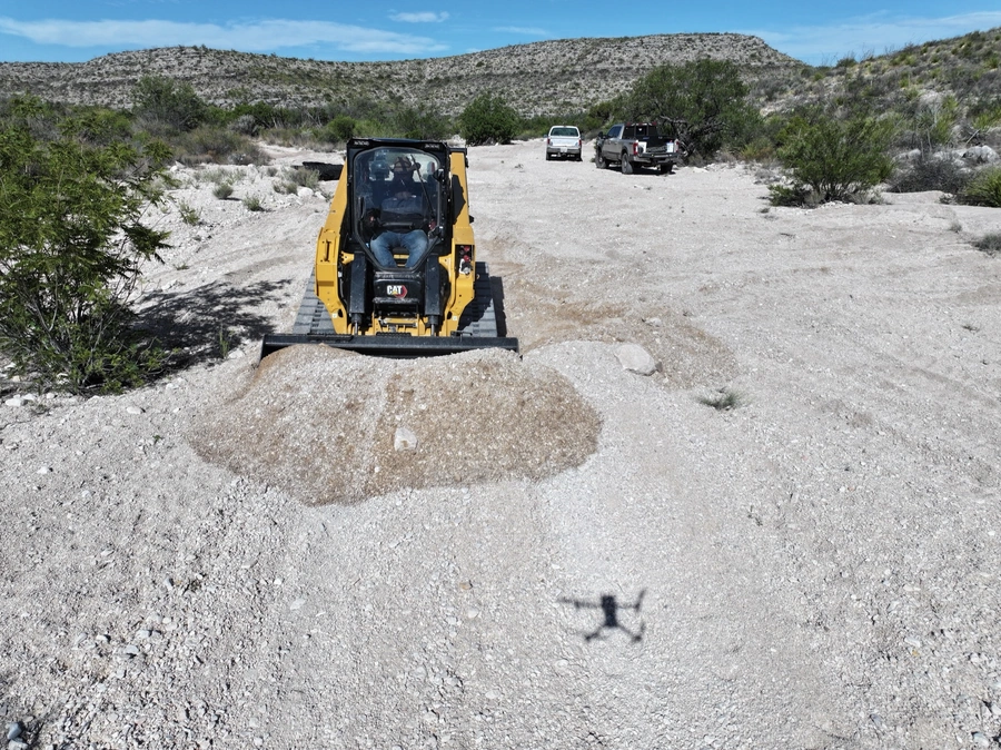 A yellow Caterpillar skid steer loader on a gravel road in a remote desert landscape, with mountains and shrubs in the background and a drone's shadow visible on the ground.