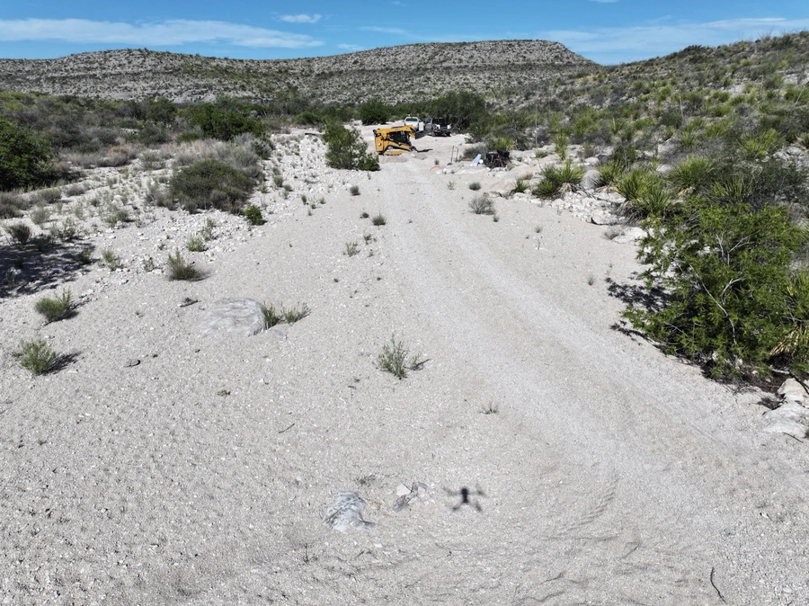 A remote desert landscape with a gravel road leading through the rocky terrain, surrounded by rugged hills and arid vegetation. A yellow construction vehicle can be seen in the distance, suggesting ongoing land improvement or site work in the area.