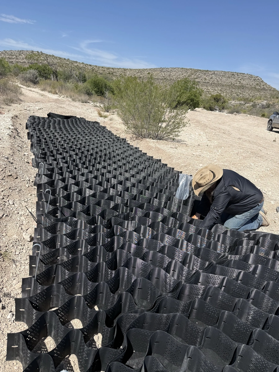 A worker is using a mat made of interlocking plastic triangles to stabilize and reinforce a sandy, rocky desert landscape.