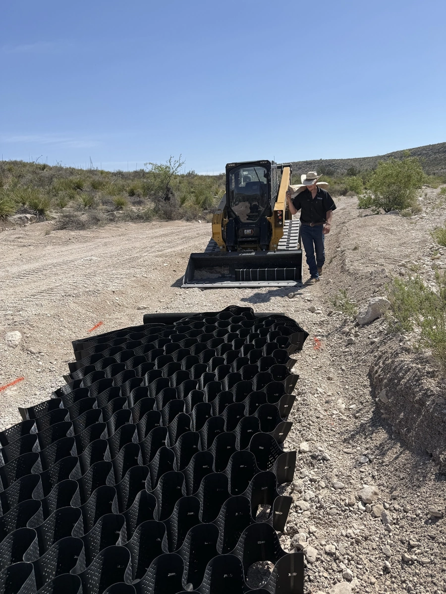 A construction worker in a Caterpillar skid steer loader operating on a dirt road in a desert landscape with scrubby vegetation and a bright blue sky.