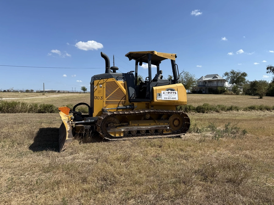 A yellow Caterpillar excavator or bulldozer machine with the A.M. Pitts Construction logo, working in a grassy field with a house visible in the background.