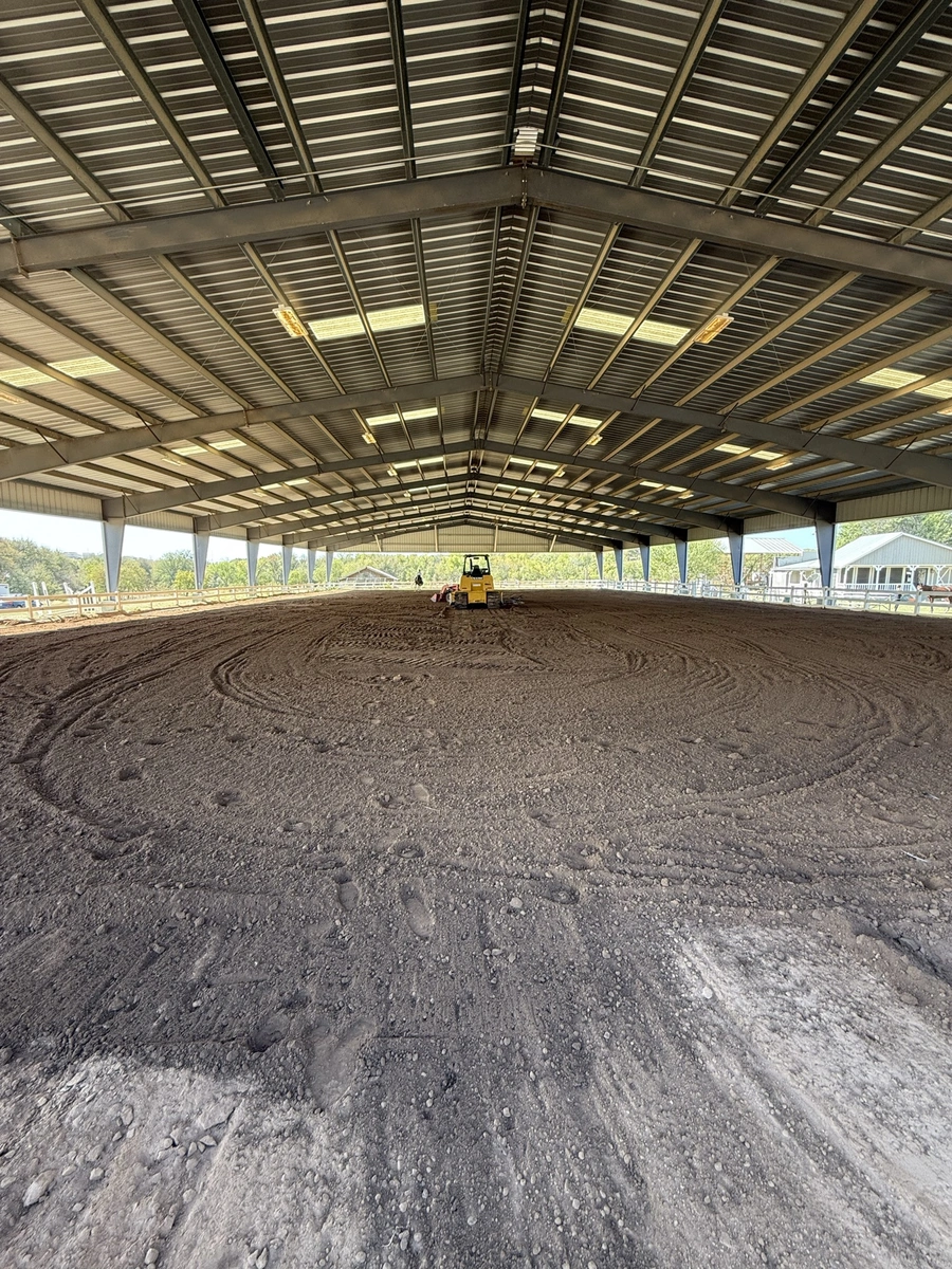 A large metal-frame building with a raised dirt floor, likely used for equestrian or agricultural purposes. A small yellow construction vehicle is visible on the floor, indicating ongoing work or preparation.