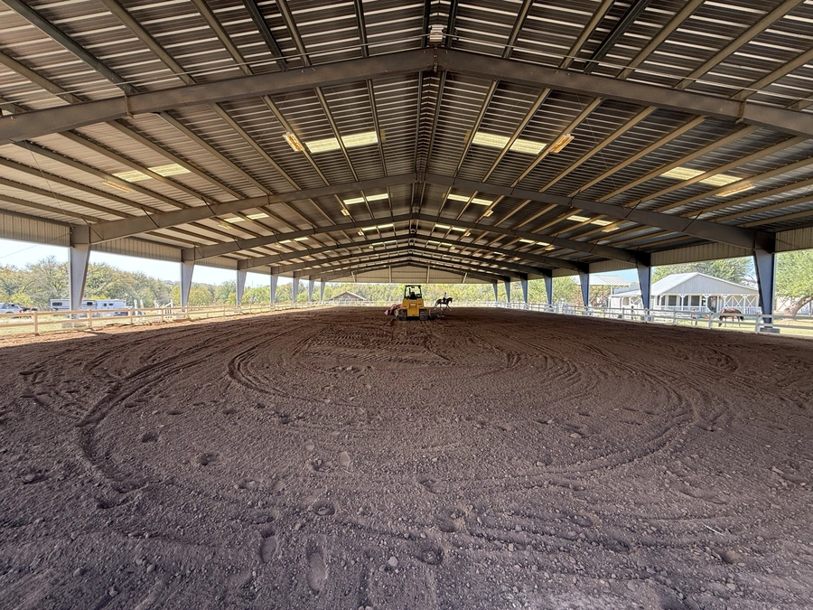 An image of a large indoor equestrian arena with a dirt floor and a tractor working on the surface. The arena has a high, vaulted roof with metal beams supporting it.