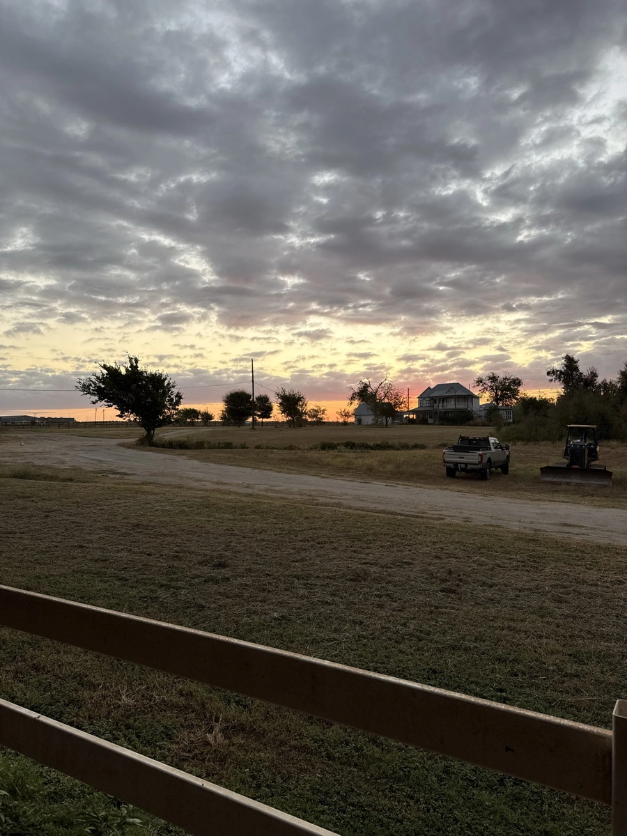 A scenic rural landscape at sunset, with a dirt road, a tractor, and a house in the background. The sky is filled with dramatic gray clouds and hues of yellow and orange.