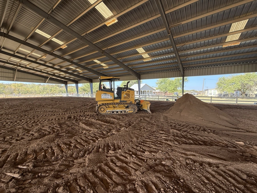 A bulldozer is leveling the ground inside a covered arena, creating a smooth surface for an equestrian facility. Piles of dirt are visible, indicating ongoing construction work.