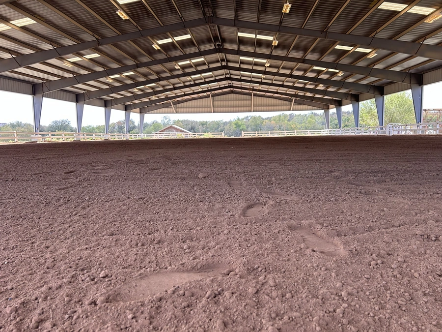 A spacious indoor arena with a large dirt floor surrounded by wooden fencing and a wooden roof structure. The arena appears to be part of a ranch or equestrian facility, with the natural surroundings visible through the open sides of the building.