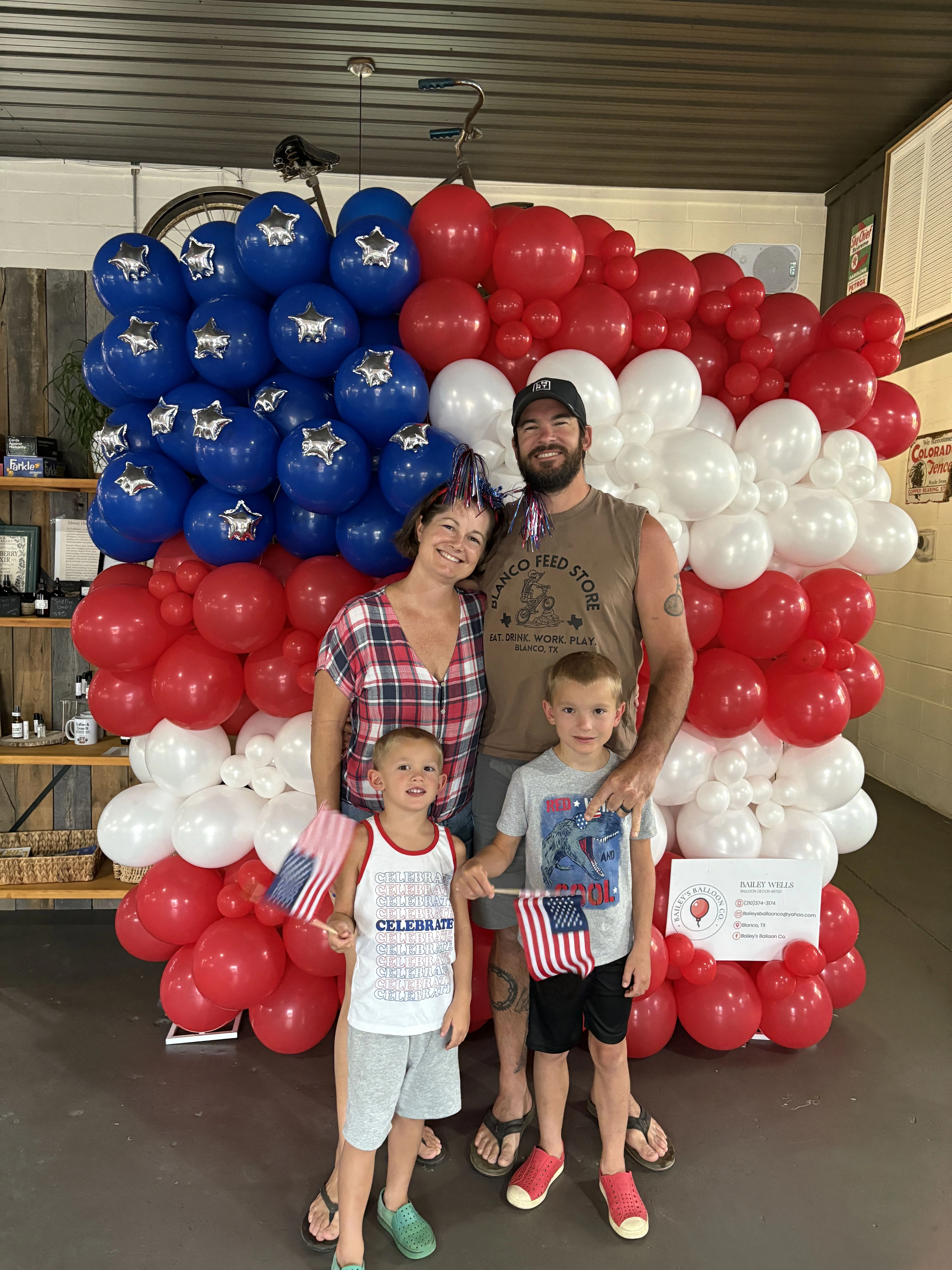 The Ketay family, the owners of the Blanco Feed Store, standing in front of a balloon mural of the US flag.