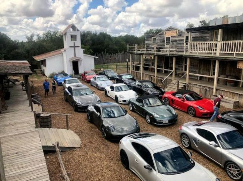 Overhead view of a car dealership lot with various makes and models of cars parked in rows. The lot includes a small building, likely an office or showroom, and the area is surrounded by trees and cloudy sky.