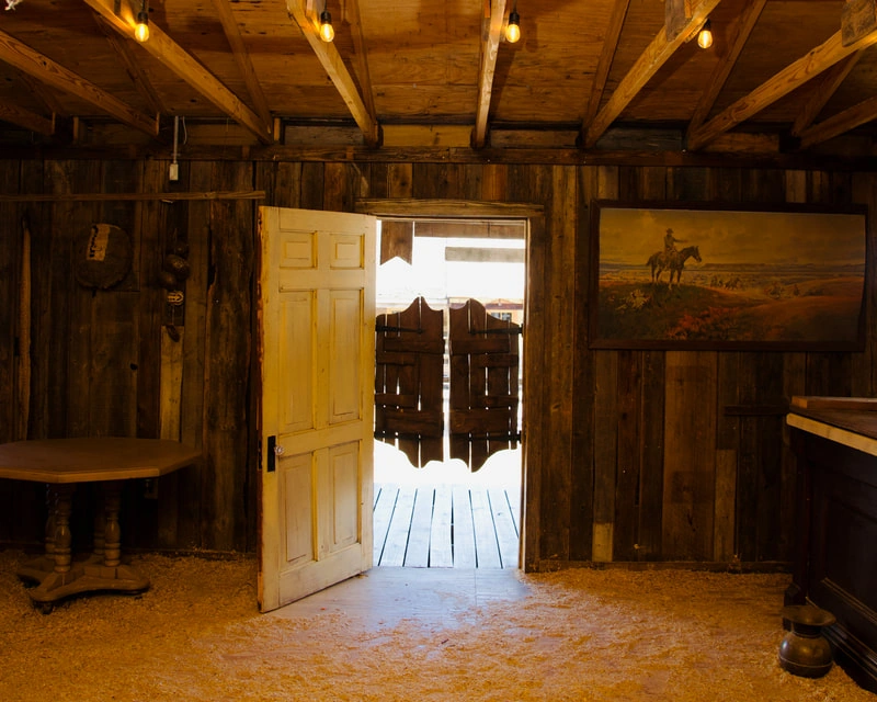 An old, rustic wooden cabin interior with a partially open wooden door leading to a snowy outdoor scene.