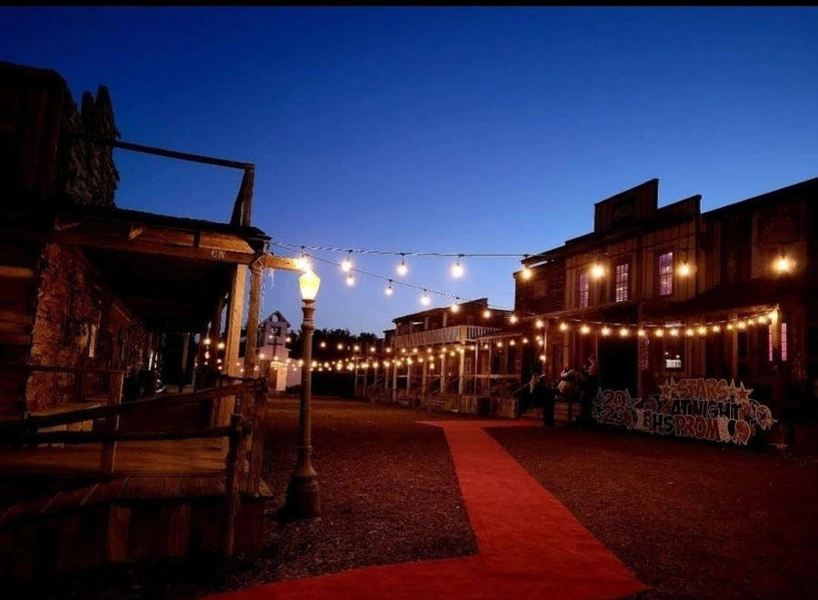 An outdoor nighttime scene with a string of festive lights strung overhead, illuminating a paved walkway lined with small businesses and restaurants. Several people can be seen walking along the path under the warm glow of the lighting.