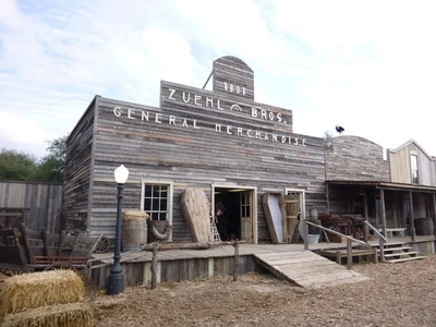 An old, weathered wooden building with a sign that says "General Merchandise" on the front. There are piles of hay or straw in front of the building, suggesting a rural or frontier setting.