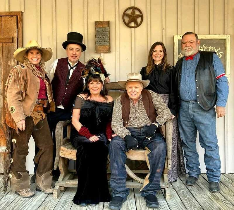 A group portrait of a diverse family or friends posing on the porch of an old wooden building. They are dressed in period costumes suggesting an old west or frontier theme, with hats, dresses, and Western-style outfits. The group includes a mix of men, women, and children of various ages.