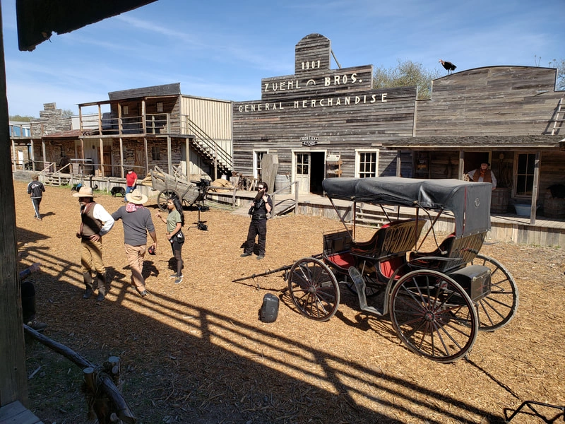 A bustling Wild West town scene with wooden buildings, a horse-drawn carriage, and people walking on a dirt road. The image evokes the frontier spirit and nostalgia of the American Old West.