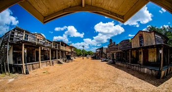 A picturesque old western town with wooden buildings and a dirt road, framed by a wooden arch. The sky is blue with puffy white clouds, creating a scenic and nostalgic atmosphere.