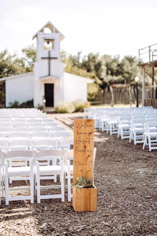 An outdoor wedding ceremony venue with a white chapel in the background, rows of white chairs set up on the ground, and a wooden sign post in the foreground with a handwritten message.