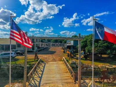 An outdoor scene with an American flag and Texas flag flying, a wooden walkway leading to a sign for Pine Moore, and a scenic landscape with trees and clouds in the background.