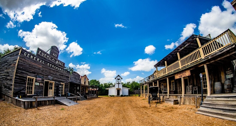 An Old West-style town with wooden buildings, dirt roads, and a statue in the center of the scene under a bright blue sky with fluffy white clouds.